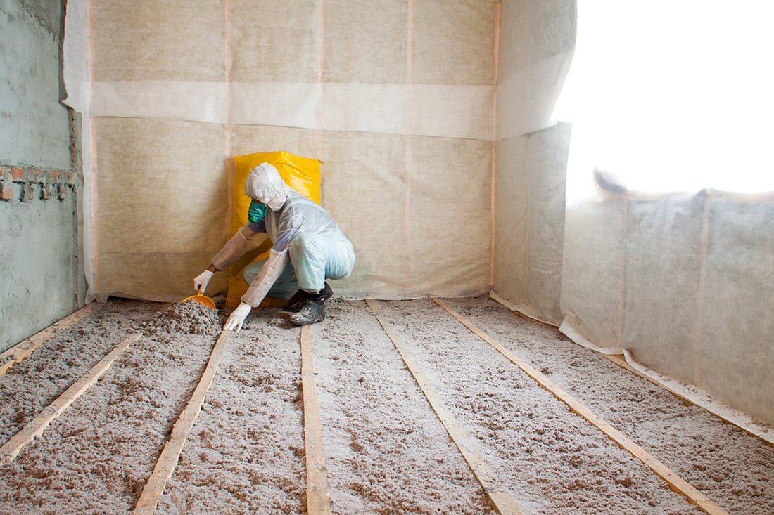 A technicians in a full body suite and dust mask installs loose fill insulation between floor joists.
