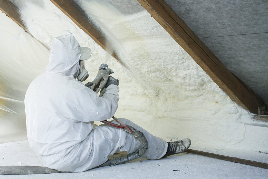 A professional in a full body suit sits in an attic an applies spray foam insulation between wood beams. 